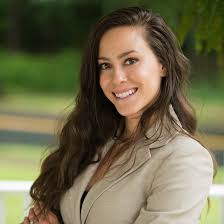 Woman in a beige blazer standing outdoors with greenery in the background
